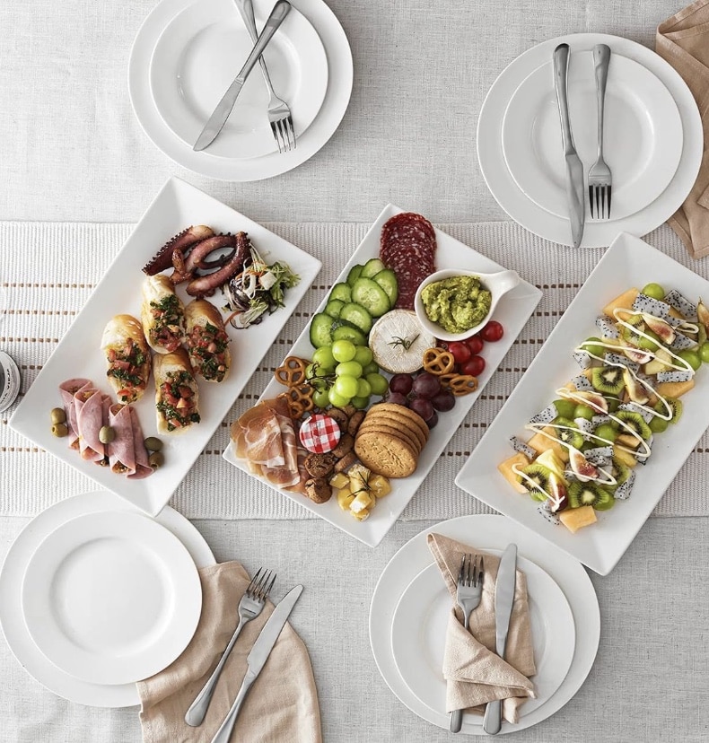 Aerial view of a dining table set with three rectangular plates. They hold an assortment of foods: bruschetta, assorted meats, cheeses, fruits, pastries, and a delightful slice inspired by a healthy lemon loaf recipe. Surrounding the plates are empty white plates, forks, knives, and beige napkins.